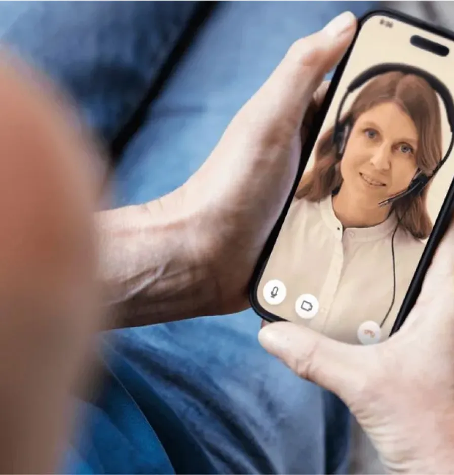 Hände halten ein Smartphone mit Videoanruf, auf dem Bildschirm lächelt eine Frau mit Headset