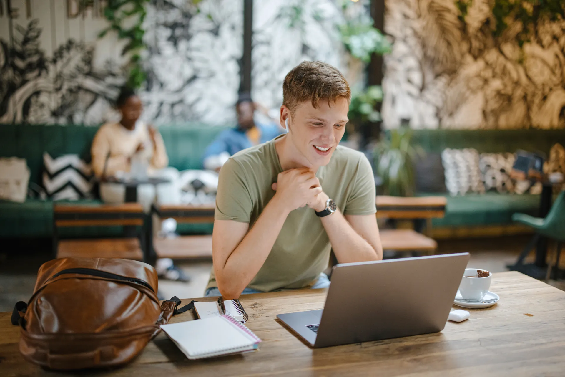 Junger Mann mit In-Ear-Kopfhörern sitzt im Café und schaut lächelnd auf seinen Laptop
